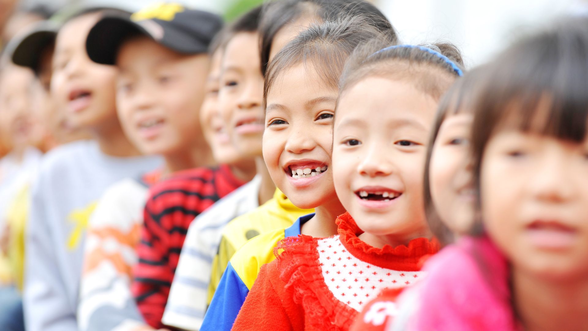 Những đứa trẻ tươi cười xếp thành hàng, mặc quần áo nhiều màu sắc.