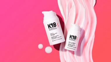 Hai chai sản phẩm chăm sóc tóc K18 màu trắng được đặt trên nền hồng cùng lớp kem xoáy mềm mại.