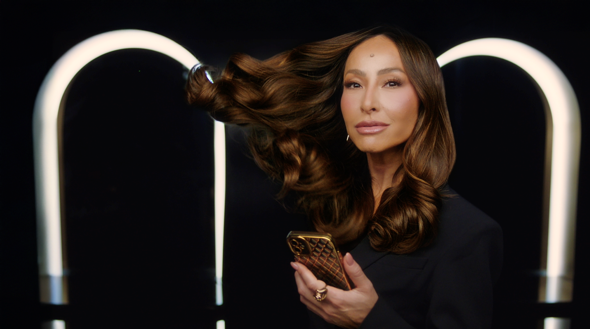 Một người phụ nữ hất mái tóc dài màu nâu của mình trong khi cầm điện thoại thông minh trước ánh đèn rực rỡ.