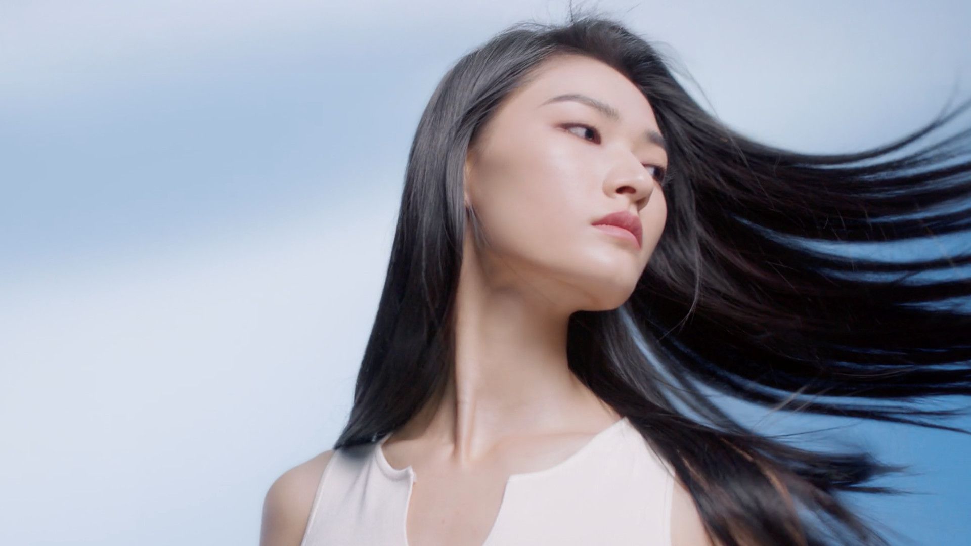  Người phụ nữ với mái tóc dài đen nhánh buông xõa sang một bên, trên nền xanh lam chuyển sắc, mặc áo không tay màu sáng.