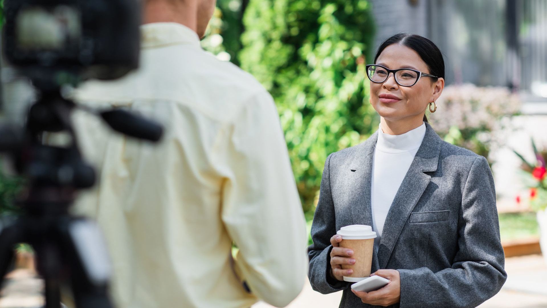 Một phụ nữ chuyên nghiệp đeo kính, tay cầm cốc cà phê, đang được phỏng vấn trước ống kính ngoài trời trong khung cảnh đô thị đầy nắng.
