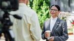Một phụ nữ chuyên nghiệp đeo kính, tay cầm cốc cà phê, đang được phỏng vấn trước ống kính ngoài trời trong khung cảnh đô thị đầy nắng.