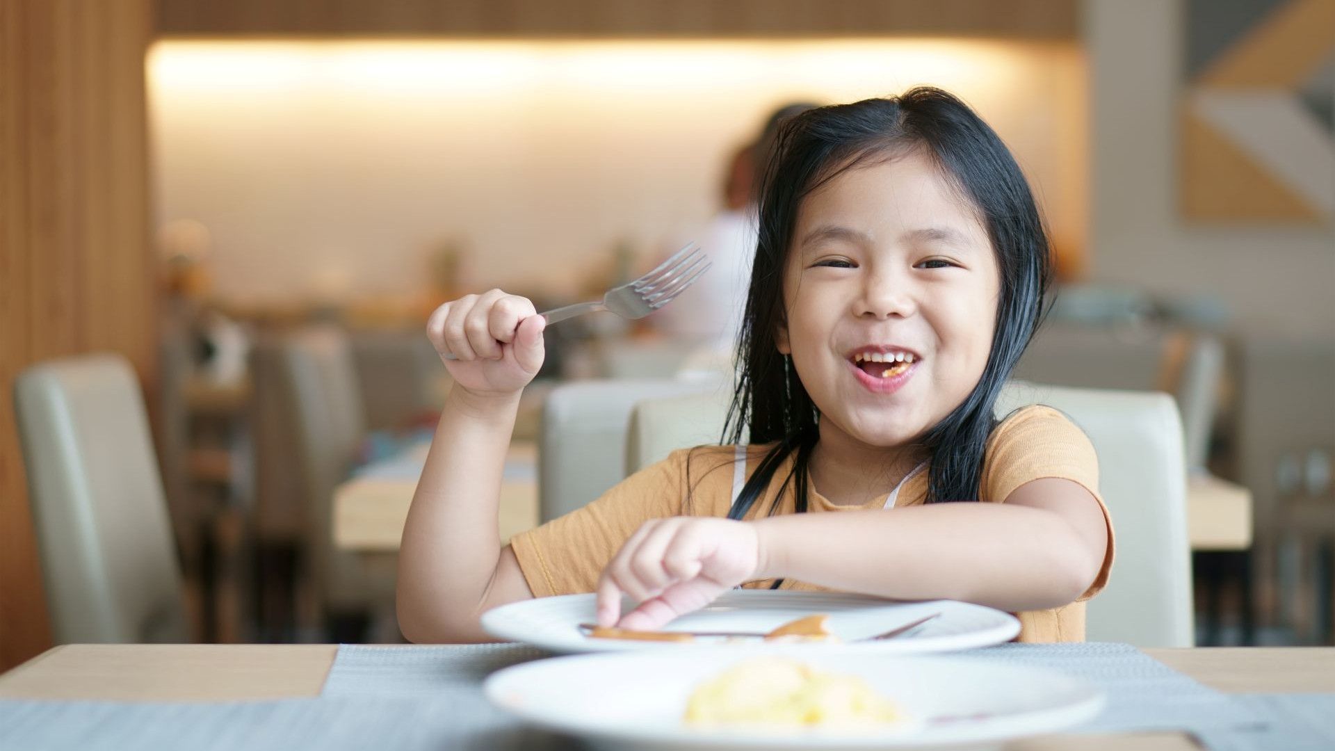 Một em bé tươi cười cầm dĩa ăn tại bàn ăn trong nhà hàng.